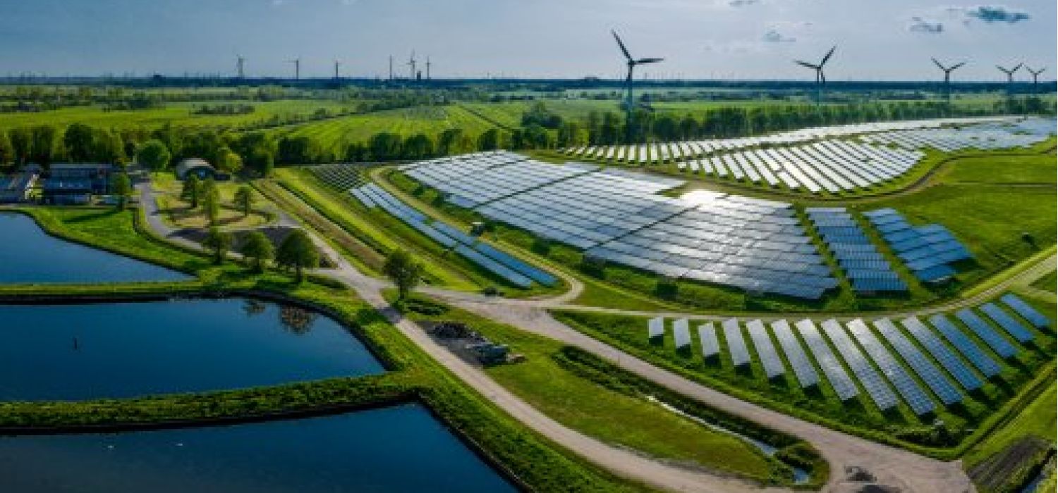 Vista aerea di un parco fotovoltaico con file di pannelli solari immerso in un paesaggio verde con laghi e turbine eoliche sullo sfondo
