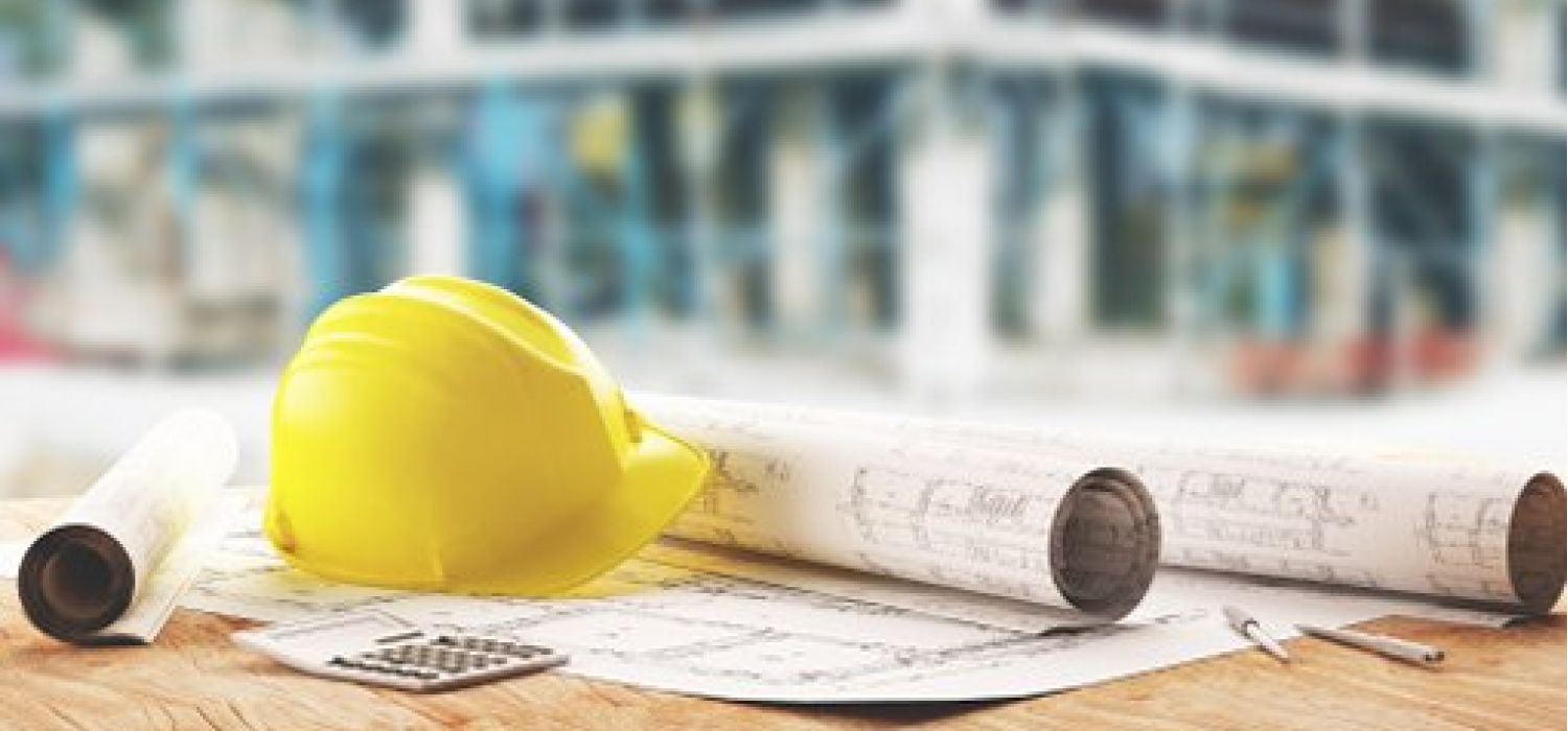 Elmetto giallo, rotoli di progetti e calcolatrice su un tavolo da lavoro con cantiere edile sfocato sullo sfondo