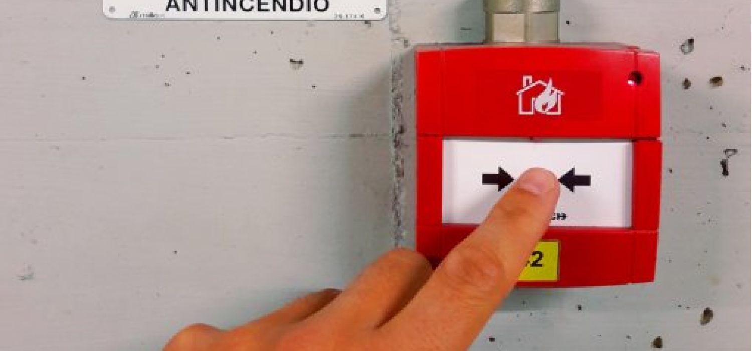 Mano che preme un pulsante di allarme antincendio rosso a parete con targa identificativa