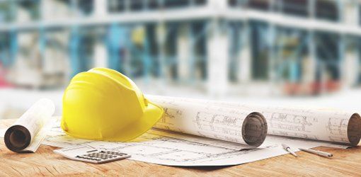 Elmetto giallo, rotoli di progetti e calcolatrice su un tavolo da lavoro con cantiere edile sfocato sullo sfondo