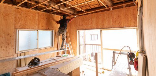 Operaio su scala che lavora al soffitto in legno di un locale in ristrutturazione, con pareti in compensato e finestre che danno sull'esterno