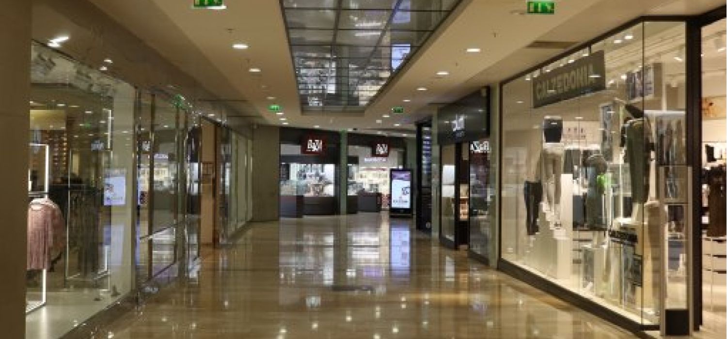 Corridoio interno di un centro commerciale vuoto con negozi di abbigliamento e vetrine illuminate su pavimento in marmo lucido