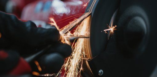 Mano con guanto nero che affila un oggetto metallico su una mola, con scintille arancioni in primo piano