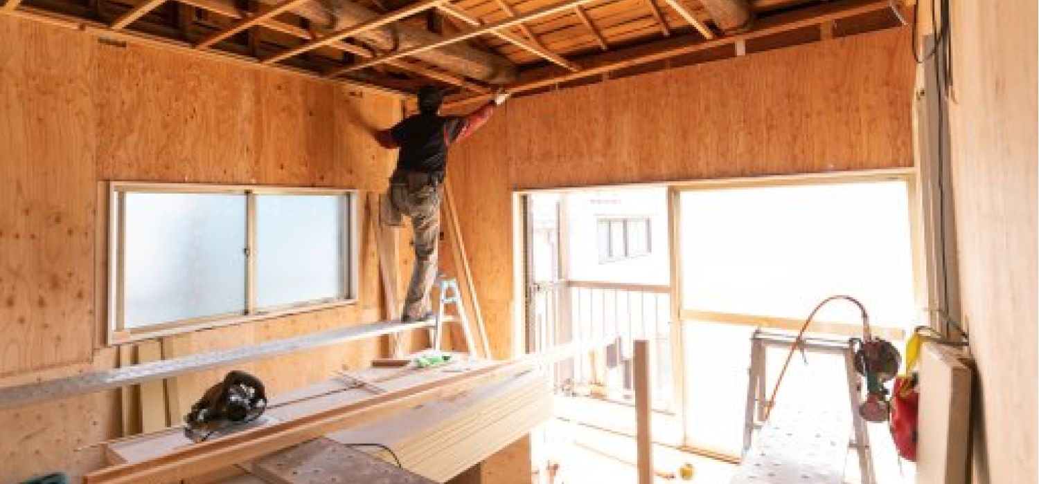 Operaio su scala che lavora al soffitto in legno di un locale in ristrutturazione, con pareti in compensato e finestre che danno sull'esterno