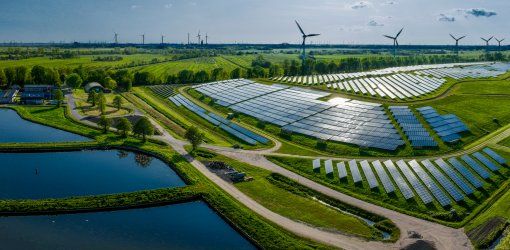 Vista aerea di un parco fotovoltaico con file di pannelli solari immerso in un paesaggio verde con laghi e turbine eoliche sullo sfondo